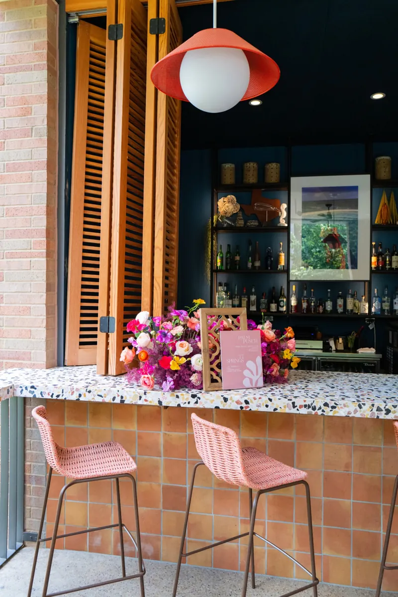 Pink bar sign and bright florals on a terrazzo bar with pink stools