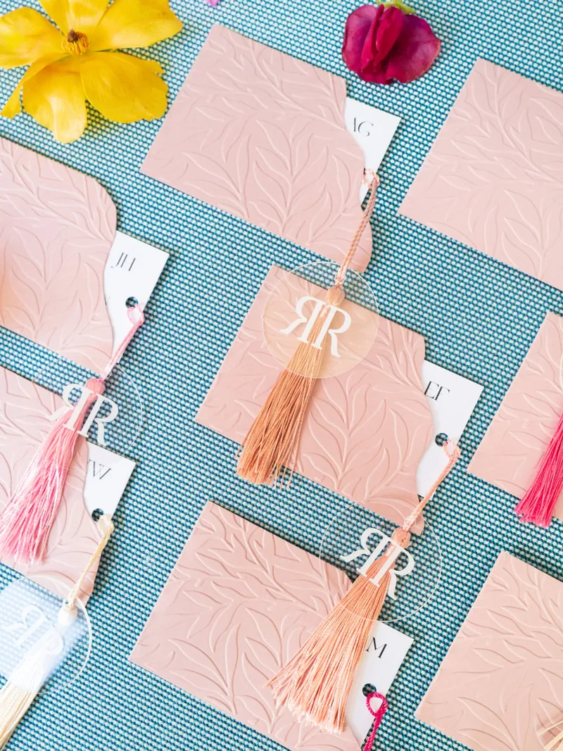 Pink leaf-embossed cards with acrylic monogram tags and coral tassels on a teal tablecloth