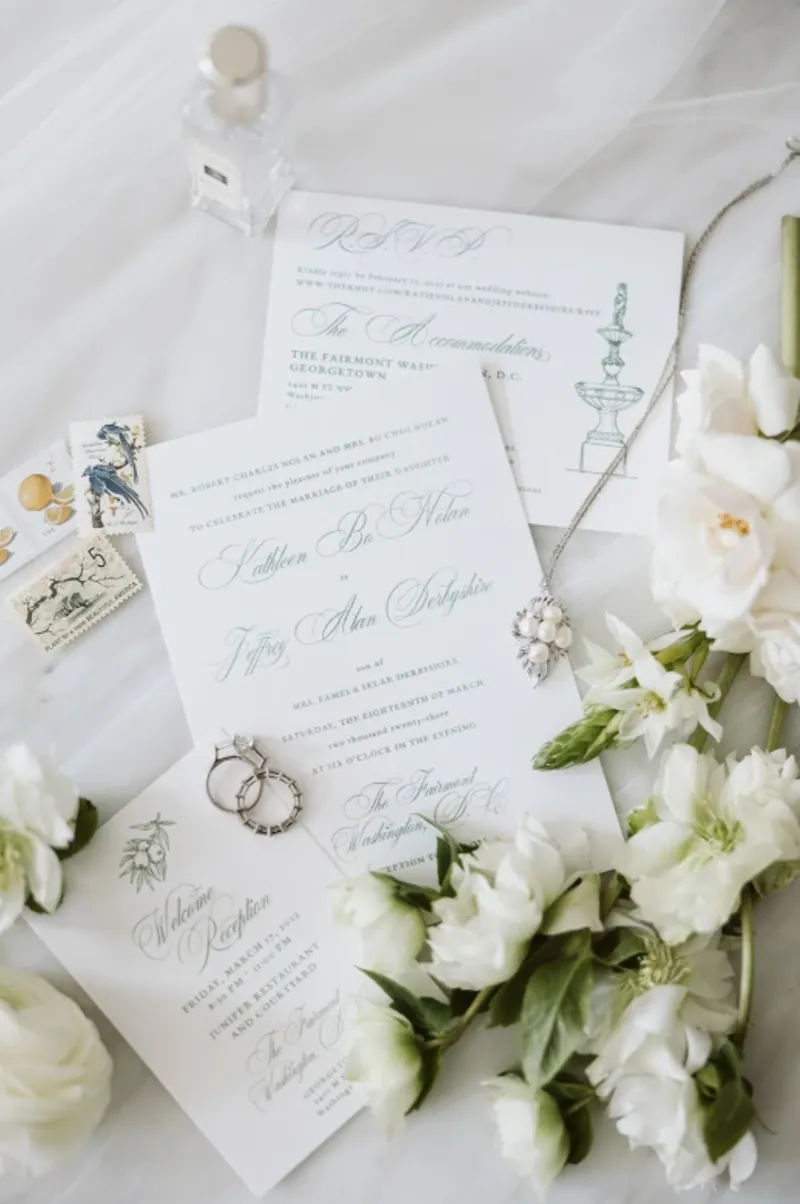 Classic white invitation suite with script lettering, vintage stamps, white florals, and rings